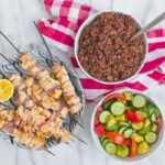 6 skewers of chicken kabobs on a marble table top next to a bowl of quinoa and a bowl of sliced cucumber and tomato salad