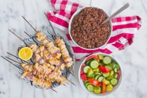6 skewers of chicken kabobs on a marble table top next to a bowl of quinoa and a bowl of sliced cucumber and tomato salad