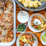 Chilaquiles, traditional Mexican breakfast dish made from crisp corn tortillas sautéed with fresh salsa, then topped with eggs, cotija cheese and onions, in a serving dish with plates set out on top of orange fabric napkins.
