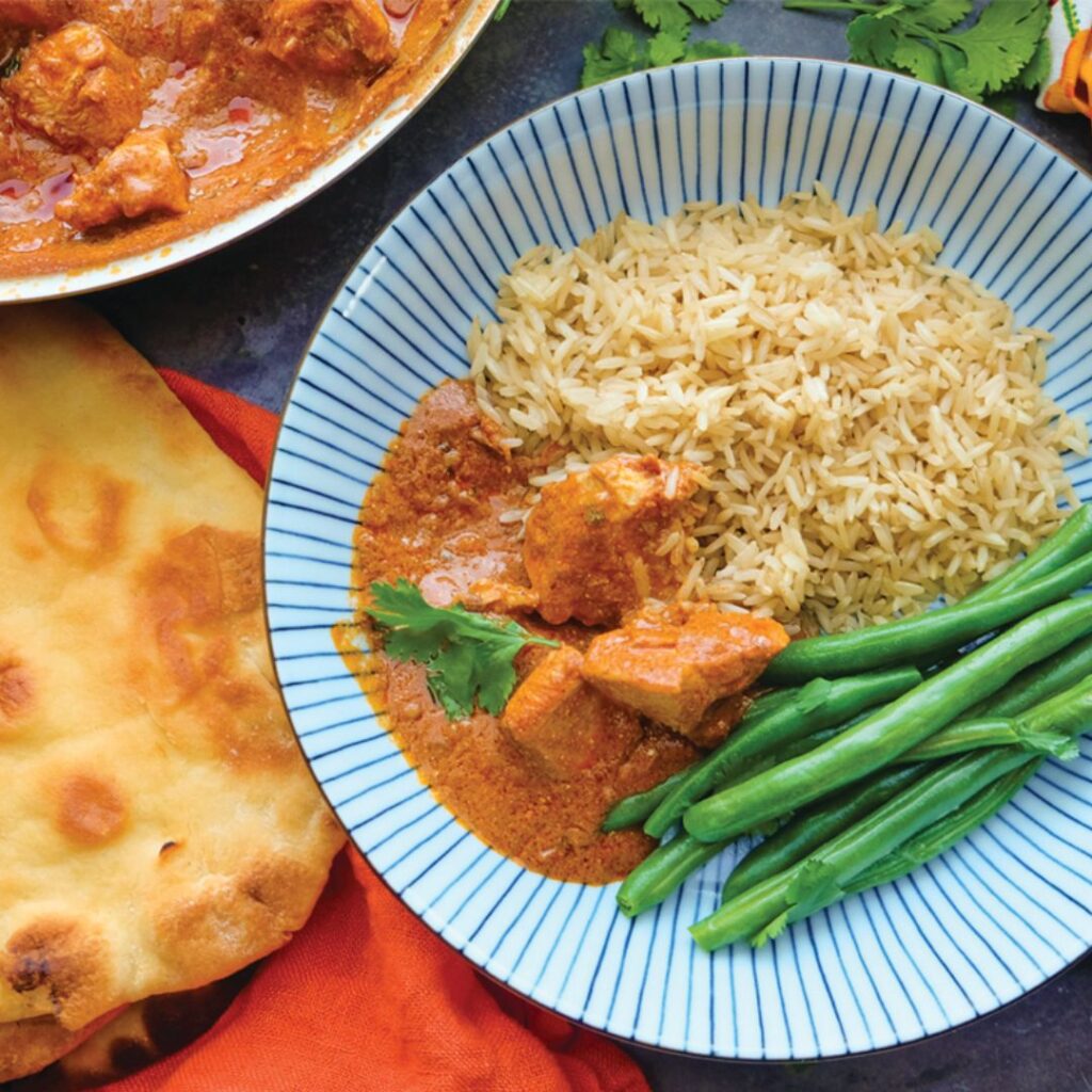 Chicken Tikka Masala served on a blue striped plate with brown rice and haricot vert, naan bread sitting to the side of the plate
