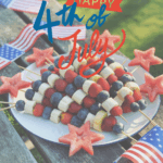 A platter of fresh fruit on a picnic table. The platter has skewers of blueberries, sliced strawberries, and sliced bananas alternating in a red, white, and blue pattern. Each skewer is topped with a watermelon shaped in a star cut-out. The scene is festive, decorated for the fourth of July holiday.