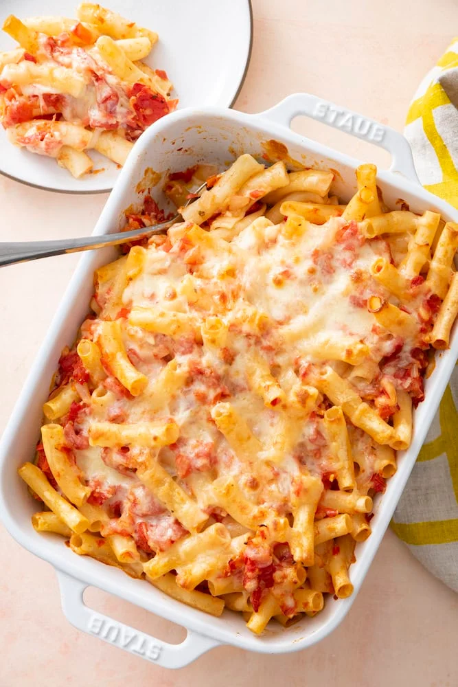 Baked ziti with melted, browned mozzarella in a white baking dish, with a serving on a plate