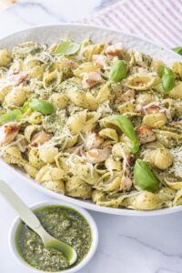 Shell pasta tossed with basil pesto, chopped chicken and Parmesan in a white bowl, with a small dish of pesto on the side
