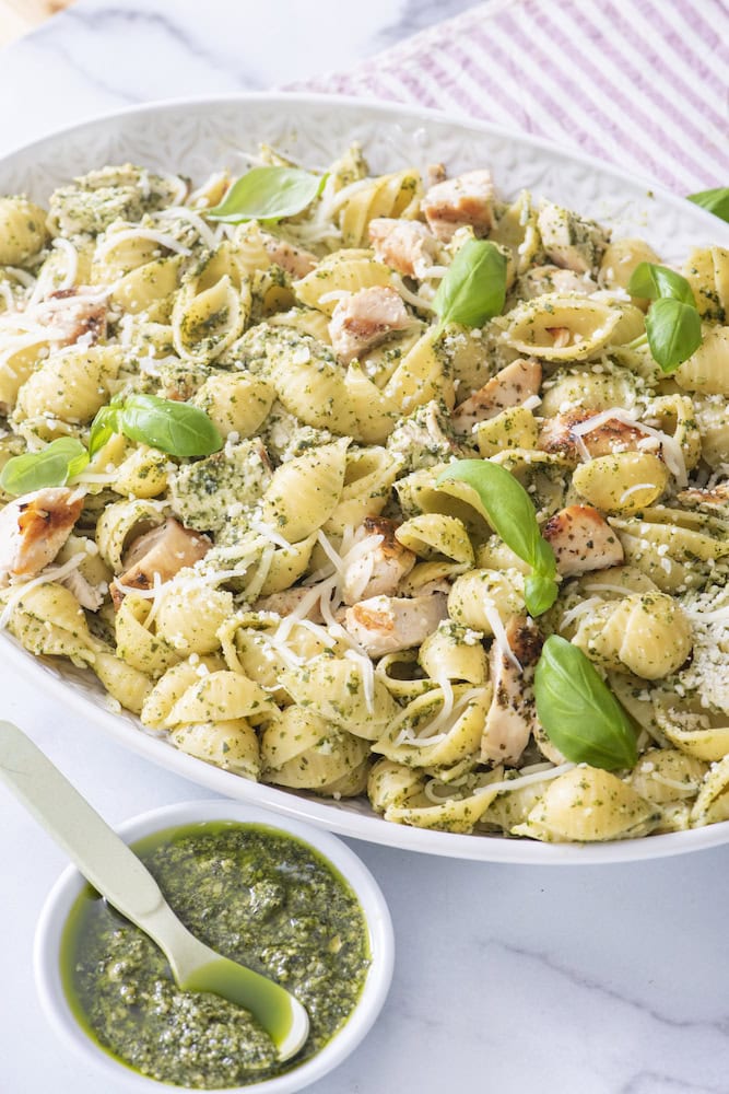 Shell pasta tossed with basil pesto, chopped chicken and Parmesan in a white bowl, with a small dish of pesto on the side