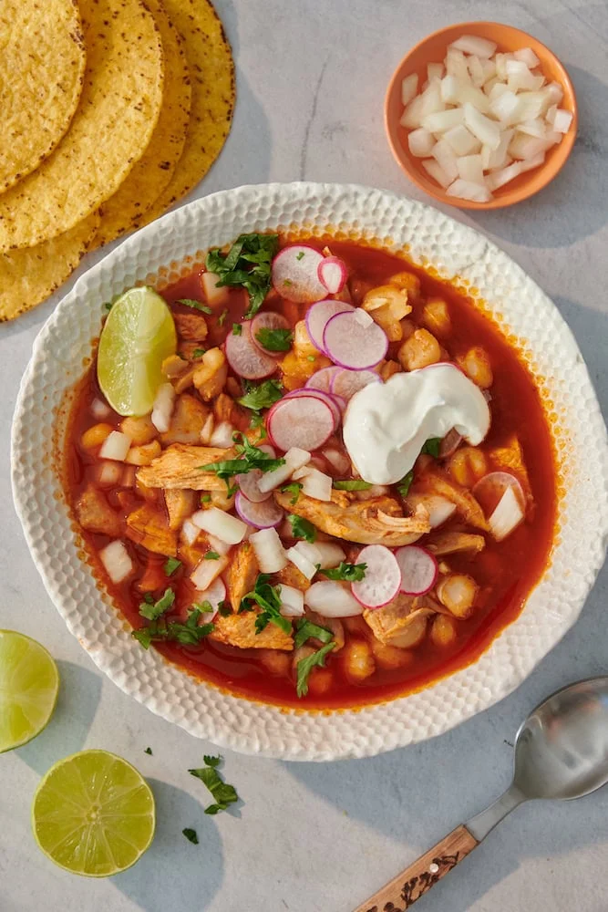 Bowl of red chicken pozole topped with radishes, onion and lime