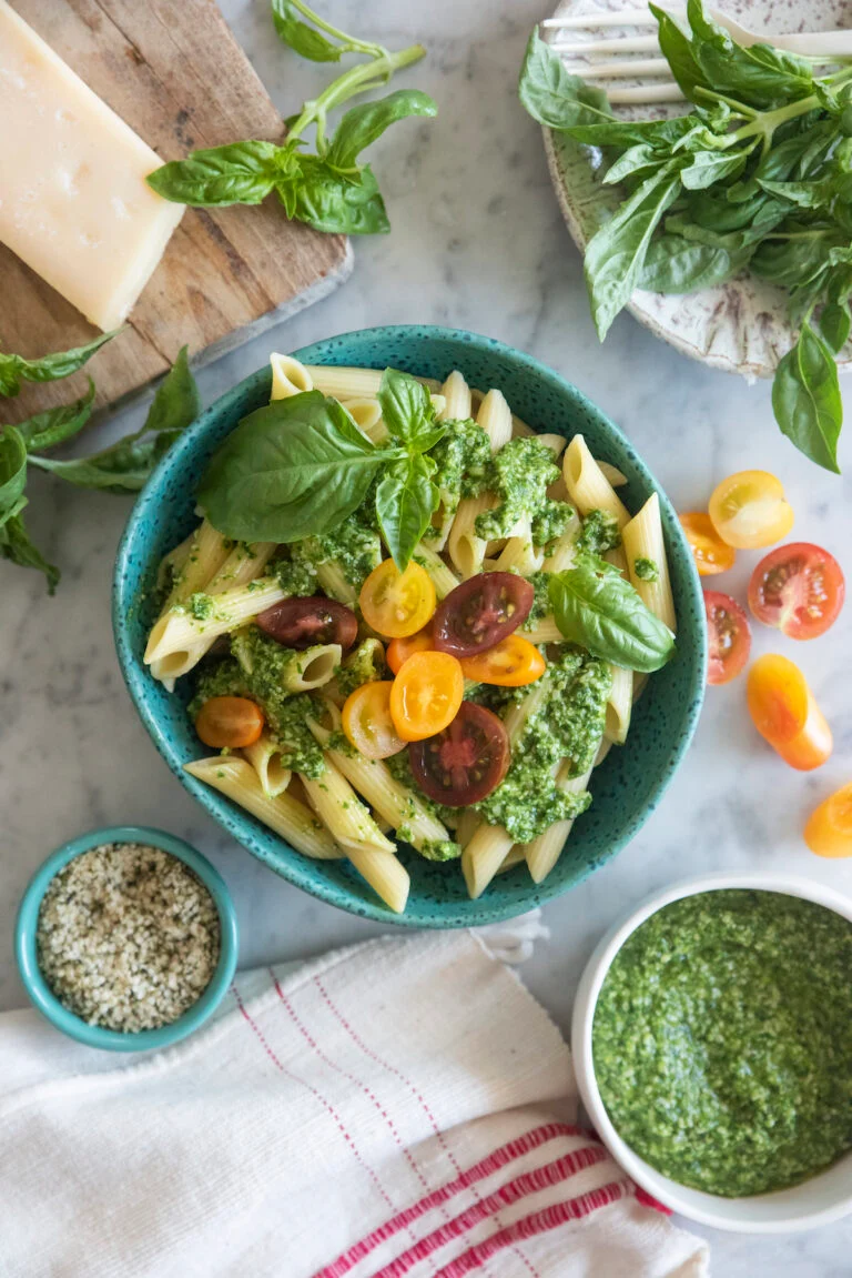 Penne tossed with hemp seed pesto, fresh basil and cherry tomatoes in a teal bowl, with a dish of extra pesto and hemp seeds