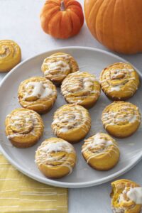 Glazed pumpkin cinnamon rolls on a plate next to small pumpkins