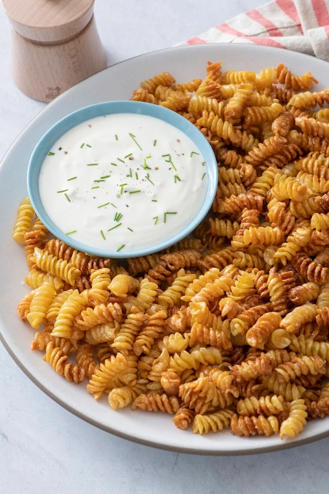 Crispy air-fried rotini pasta chips on a platter with a bowl of herby yogurt dip
