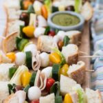 Sandwich skewers with bread, ham, cheese, cucumber and tomatoes beside a bowl of pesto