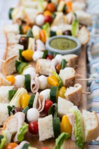 Sandwich skewers with bread, ham, cheese, cucumber and tomatoes beside a bowl of pesto