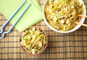 Two bowls of sesame noodles with tofu, mushrooms and scallions on a bamboo mat, with chopsticks