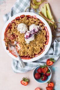 Strawberry rhubarb crumble in a baking dish topped with scoops of ice cream, with fresh strawberries