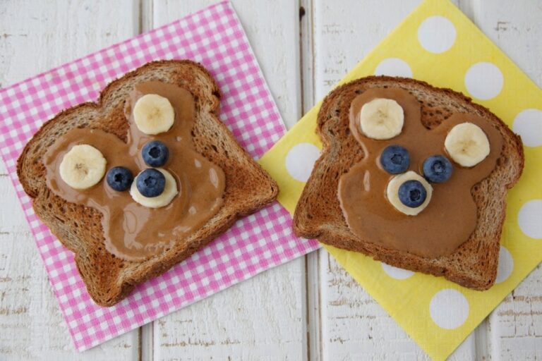 Two slices of teddy bear toast with peanut butter, banana ears and blueberry eyes