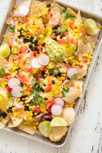Sheet pan of tortilla chips topped with cheese sauce, black beans, corn, radishes, pickled onions, jalapeños, guacamole and lime wedges