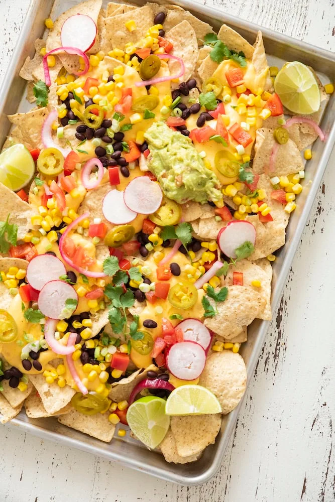Sheet pan of tortilla chips topped with cheese sauce, black beans, corn, radishes, pickled onions, jalapeños, guacamole and lime wedges