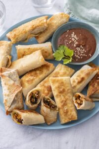 Pan-fried vegetarian spring rolls, some cut open to show the vegetable filling, on a blue plate with dipping sauce