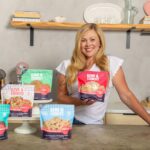 Ashley Cameron standing in a kitchen with exposed wooden shelves showing off her bags of Love & Cookies