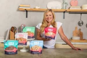 Ashley Cameron standing in a kitchen with exposed wooden shelves showing off her bags of Love & Cookies