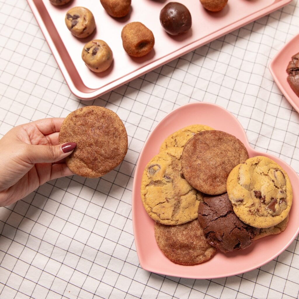a pink heart shaped plate layered with a variety of cookies, with a hand holding a cookie over grid print table cloth