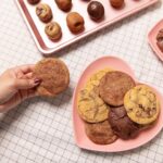 a pink heart shaped plate layered with a variety of cookies, with a hand holding a cookie over grid print table cloth