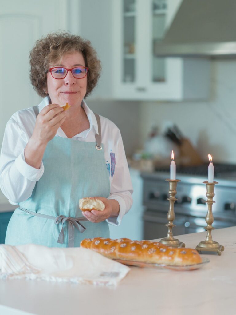 A caucasian Jewish woman wearing a blue apron stands behind a white countertop with two white Shabbat candles and a challah bread