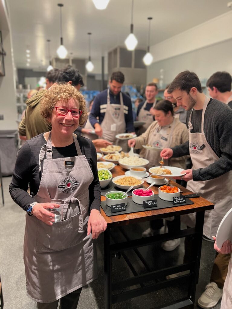 White woman stands in front of a counter in a commercial kitchen with a room full of people attending a cooking class