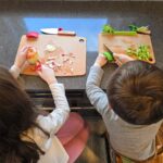Two young children, visible only from the back of their heads, sit at a kitchen counter in front of cutting boards peeling and cutting an apple each