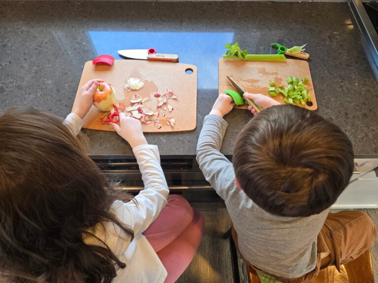 Two young children, visible only from the back of their heads, sit at a kitchen counter in front of cutting boards peeling and cutting an apple each