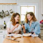 two white women sitting in a kitchen smiling and cheering with forks