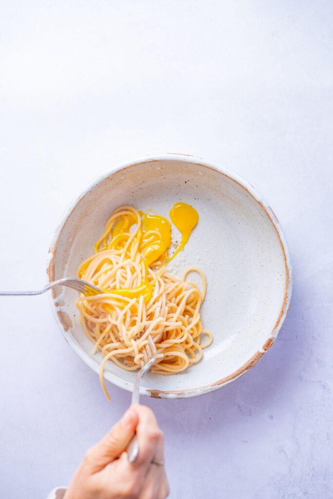 A bowl of spaghetti topped with raw egg yolks for carbonara, with a hand holding a fork