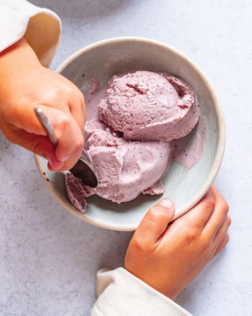 A child's hands scooping pink homemade ice cream from a bowl with a spoon