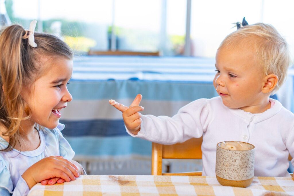 Two young children laughing together at a table, one pointing at the other