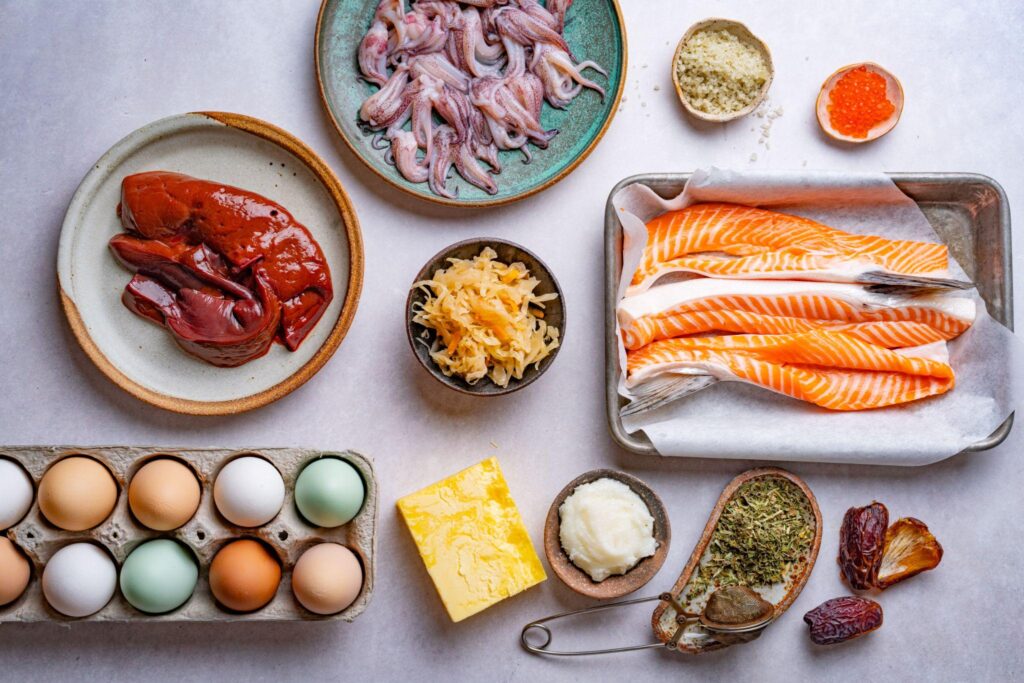 Overhead spread of nutrient-dense foods including liver, salmon fillets, eggs, sardines, fish roe, butter and dates