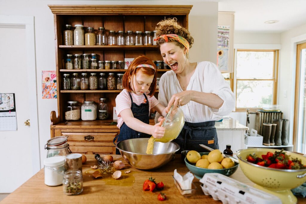 Emma Frisch, a Caucasian woman, stands smiling in her kitchen mixing ingredients into a bowl with her red-haired daughter. A bowl of strawberries, a bowl of lemons, and cracked eggshells sit on the kitchen table surrounding them.