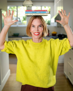 Katie Morford in a yellow sweatshirt in her kitchen, balancing her three cookbooks on her head