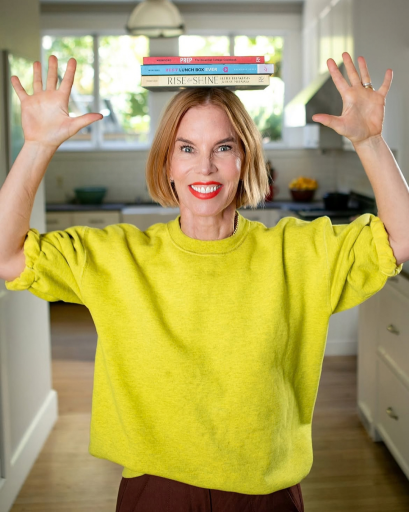 Katie Morford in a yellow sweatshirt in her kitchen, balancing her three cookbooks on her head