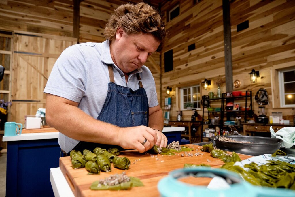 Kitchen setting, a white man stands behind a cutting board prepping stuffed grape leaves