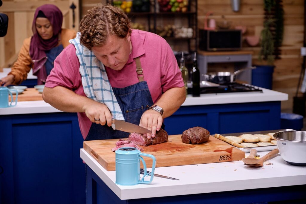 White man in a commercial kitchen on the set of Great American Recipe slicing roast meat