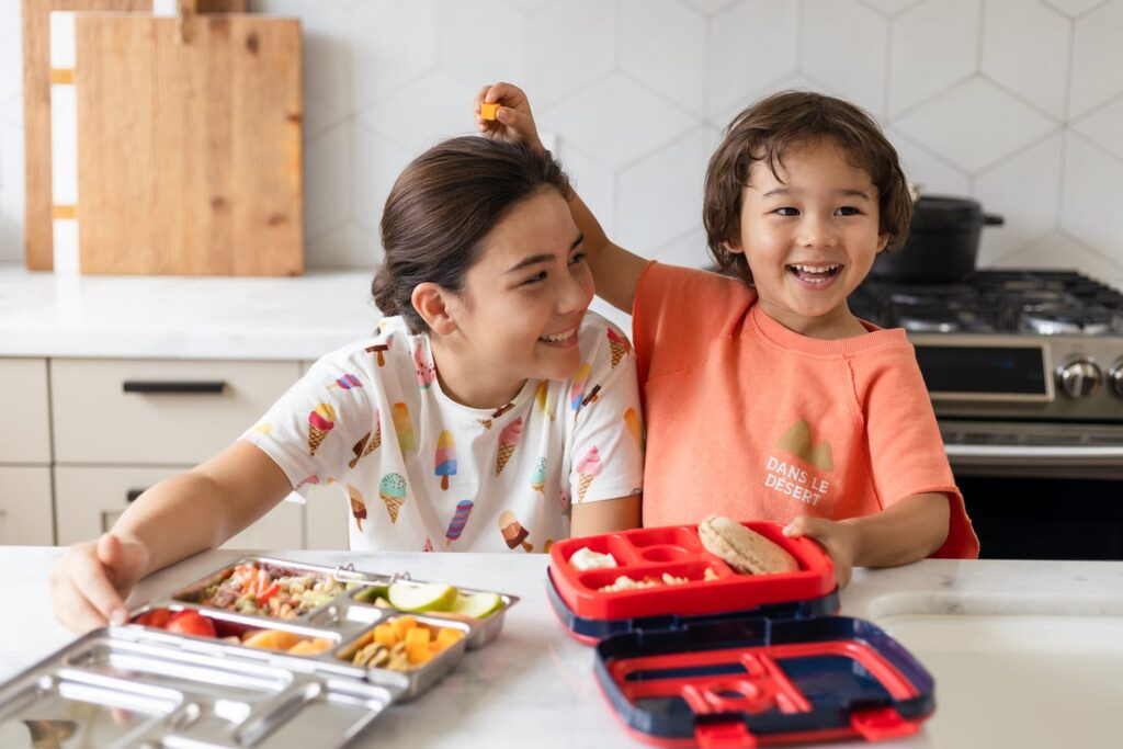 a small boy and girl in the kitchen eating from lunch boxes
