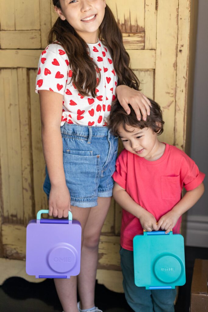 a girl in a heart t shirt and small boy in a red t shirt holding the purple and blue lunch boxes