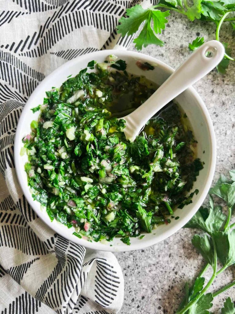 Small white bowl full of cilantro chimichurri sauce with a white spoon in it. Black and white patterned napkin and cilantro sprigs in the background.