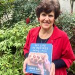 Joan Nathan in a red coat standing outside and holding her book My Life in Recipes