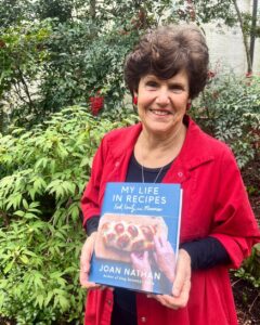 Joan Nathan in a red coat standing outside and holding her book My Life in Recipes