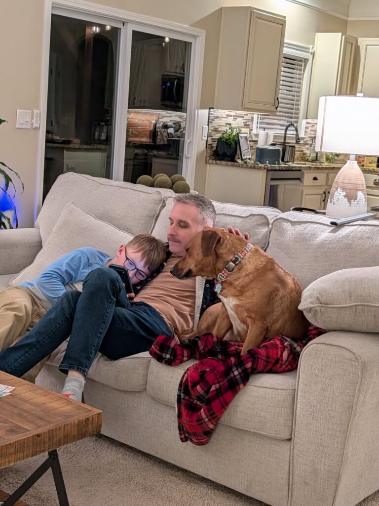 man with his son and rescue dog sitting on tan coach with red plaid blanket