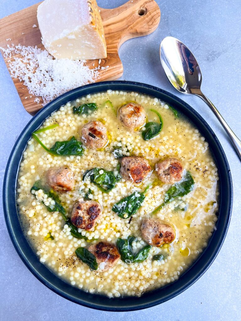 A bowl of soup with meatballs, spinach, and small pearl pasta