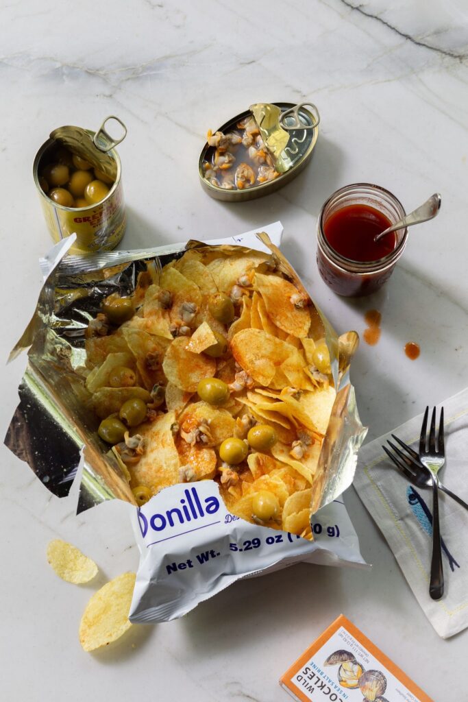 An open bag of potato chips topped with olives and hot sauce, with tins of olives and seafood