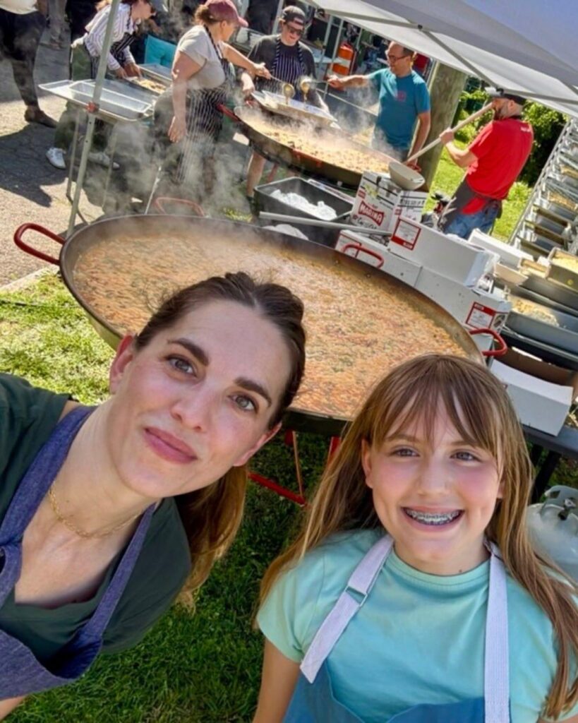 Katie Button and her daughter in aprons taking a selfie in front of a giant paella pan at an outdoor cookout