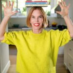 Katie Morford in a yellow sweatshirt in her kitchen, balancing her three cookbooks on her head