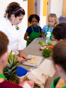A woman stands over a cutting board showing five kids how to safely use a knife