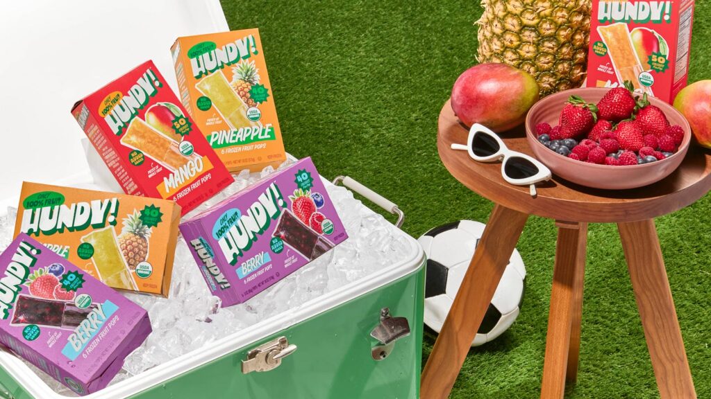 a cooler filled with ice and colorful Hundy boxes on the grass next to a small table with fruit